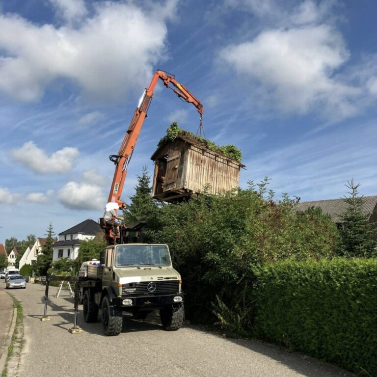 Kran hebt ein kleines Holzhäuschen über eine Hecke in einen Garten.